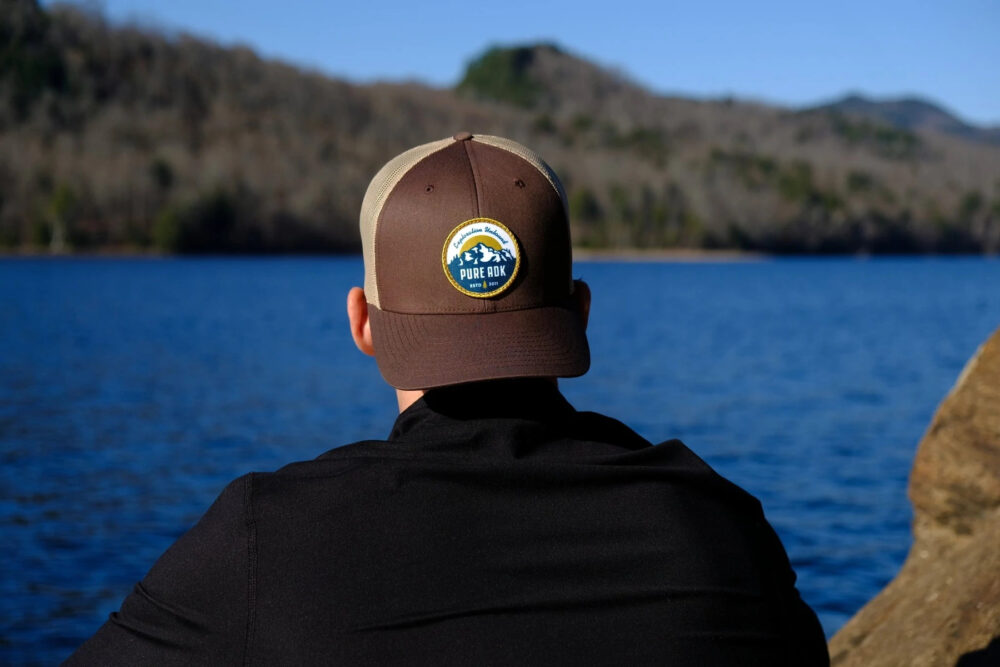 A young male sits on a shoreline with his back to the camera. He gazes off toward the distance over a beautiful lake on a sunny day. He's wearing a brown backwards Pure Adirondacks hat.