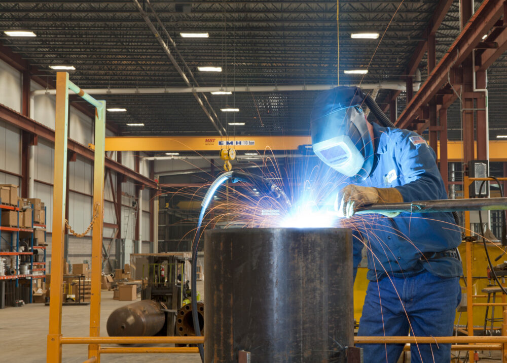 A welder in a full protective blue suit welds a piece of metal to an industrial boiler. In the distance yellow support beam can be seen in this expansive industrial warehouse.