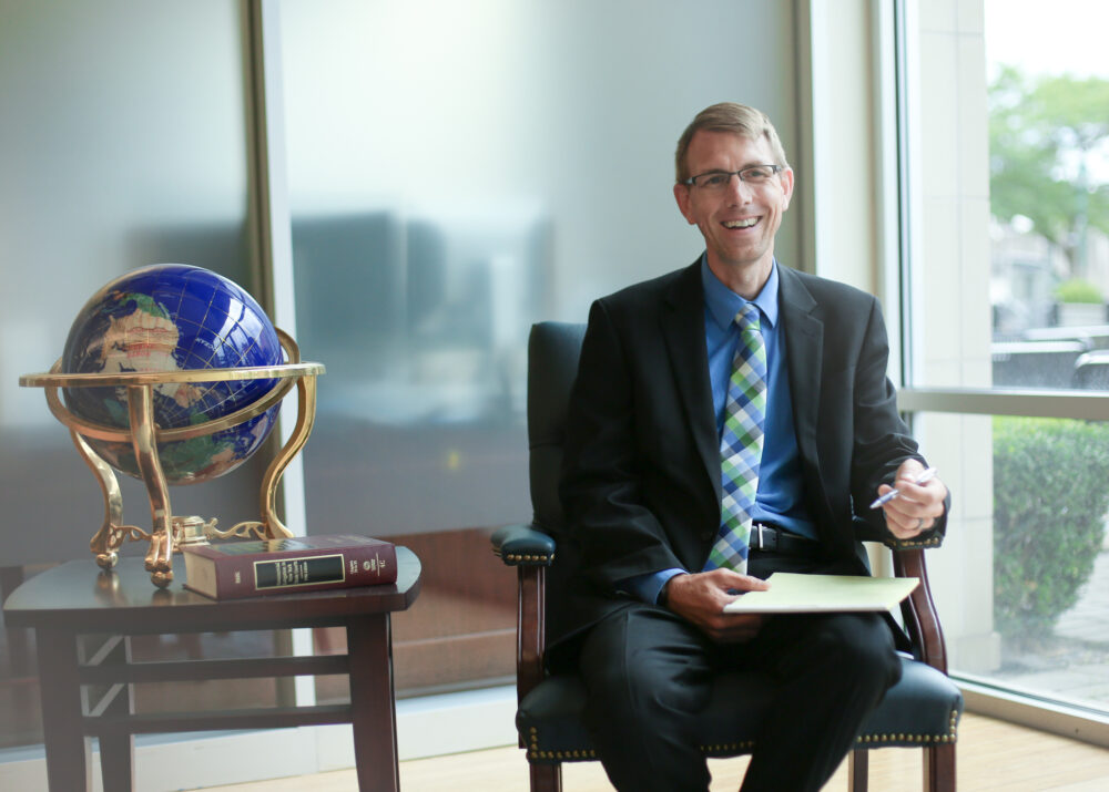 Dan Rose, a partner at Costello, Cooney & Fearon, sits smiling in a chair next to a side table holding a large golden and blue globe