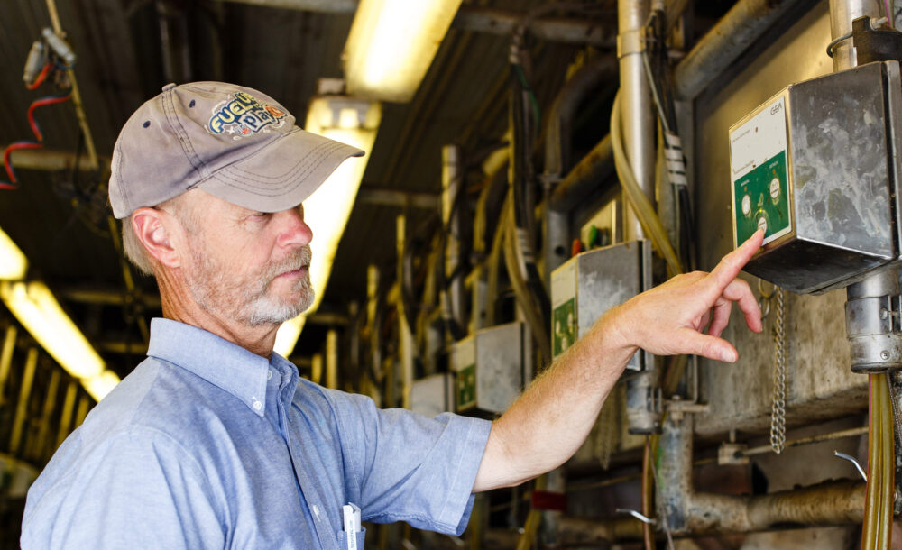 A male farmer wearing a sun bleached gray baseball cap and a blue button-down oxford shirt, holds his hand on the control mechanism in a brightly lit milking parlor.