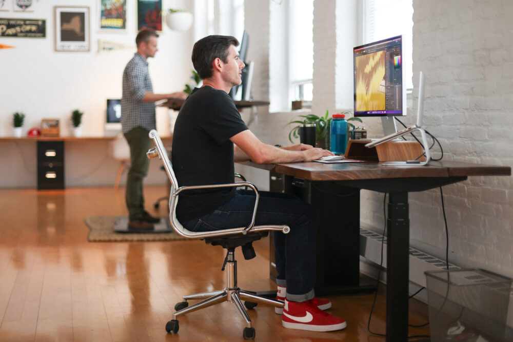 A man with red shoes sits at a desk while illustrating a poster on his computer, in the background another man works standing at a desk