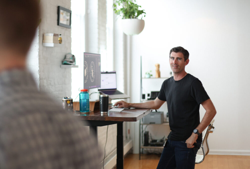 Andy Orr standing up at a desk in a brightly lit design studio with plants hanging from the ceiling and artwork on the brick walls in Syracuse, new York