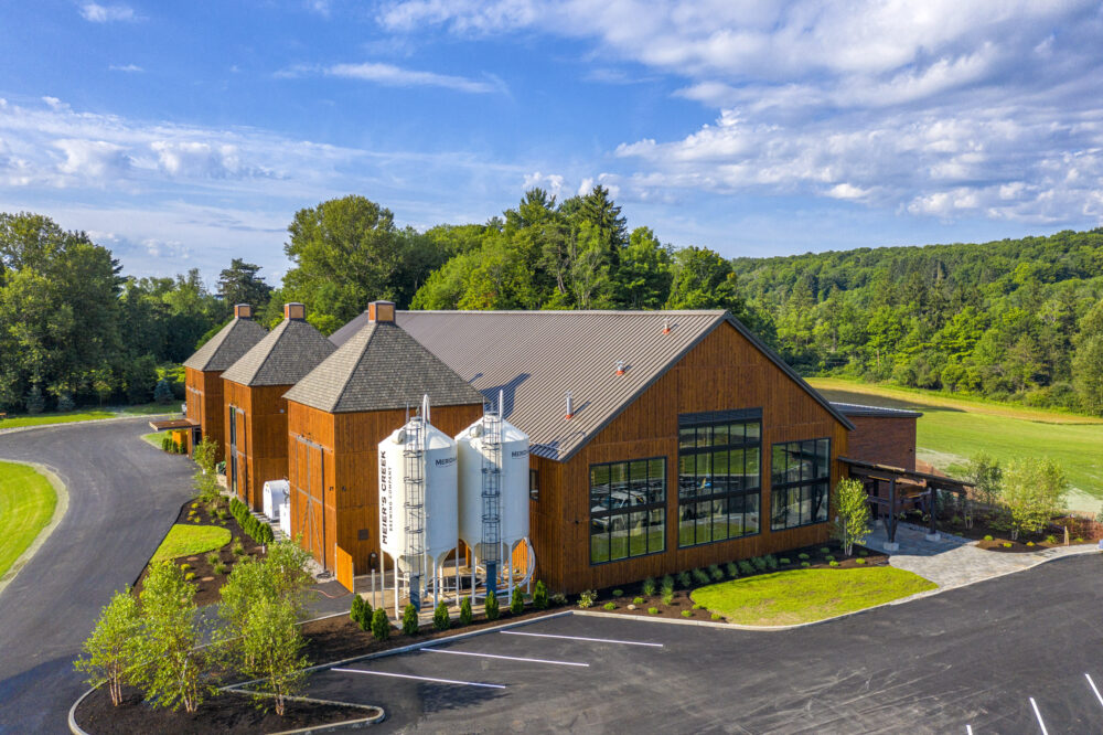 Meier's Creek Brewing Company's Cazenovia Farm Brewery location set in a beautiful clearing on a sunny day. Several white fermentation vessels are seen next to the brown exterior of the brewery.